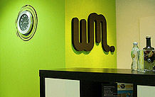 Imagen de la recepción del estudio. Aparece nuestro logotipo en negro sobre una pared pintada de verde y un expositor con algunos trabajos